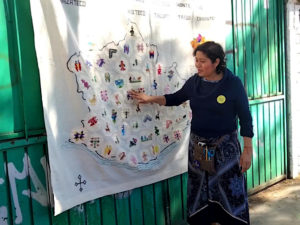 Mariana García erklärt Stickkunst vor Wandbild mit den Regionen Oaxacas zeigt