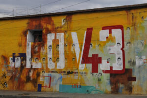 Orange-gelbe Hauserwand auf der große LOVE 43 steht. Das V und die 4 gehen ineinander über. Im Hintergrund Plakate mit Portraits der 43 Studenden, die verschwunden wurden.